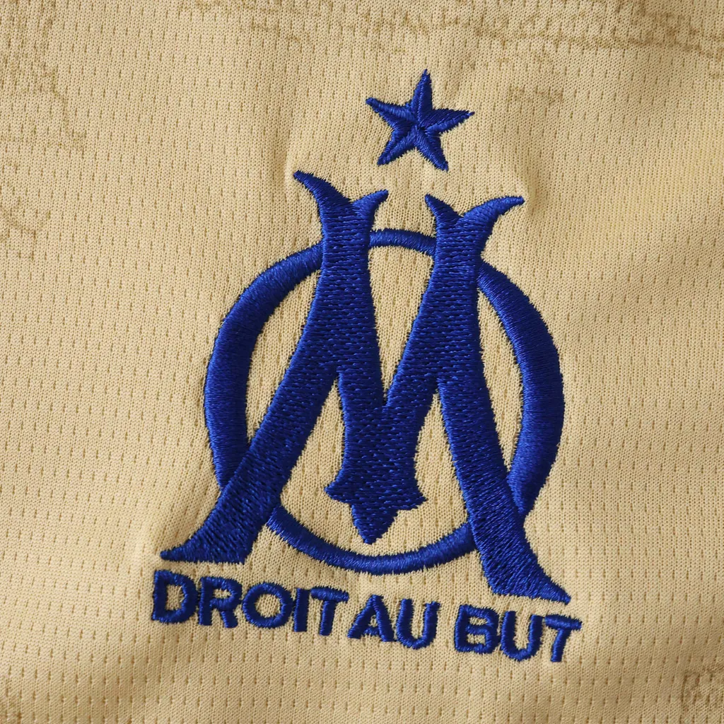 Quarta camisa do Olympique de Marselha 2025/26 Puma em tom dourado, com detalhes em azul royal, textura inspirada nos monumentos de Marselha e escudo do OM aplicado no peito.